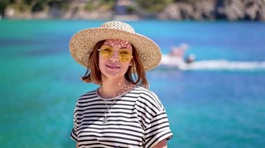 Güneş gözlüklü ve hasır şapkalı bir kadın portresi. İskelede duruyor. Tekne de yanında gidiyor. Seyahat yeri ve tatil konsepti üzerine çizgili tişörtlü bayan..