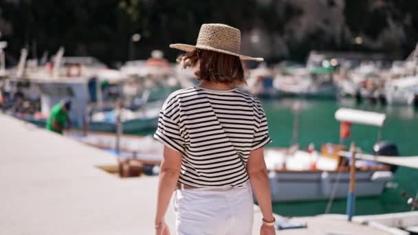 Posto barca con yacht. Giovane donna in t-shirt a righe e cappello di paglia cammina lungo la costa del sud Europa. Vacanza di lusso, viaggi, concetto di relax. — Video Stock