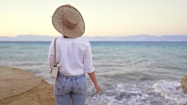 Cizí žena stojí na pláži u Středozemního Jónského moře. Dáma turista v slamáku sledování krásné modré vodní hladiny sám, příroda pozadí. Vítr, zlatá hodina. — Stock video
