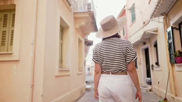 Widok z tyłu turystki w kapeluszu zwiedzanie starego europejskiego miasta na wyspie Grecja. Pani zwiedzanie lokalnej architektury śródziemnomorskiej, kultury. Dobrze się bawi włócząc się latem po mieście. — Wideo stockowe