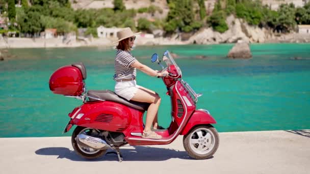 Corfou, Grèce - septembre 2021. Femme assise sur Vespa scooter moto en bonbons couleur rouge pomme, jetée au village Paleokastritsa, station de luxe. Incroyable fond d'eau de mer turquoise. — Video