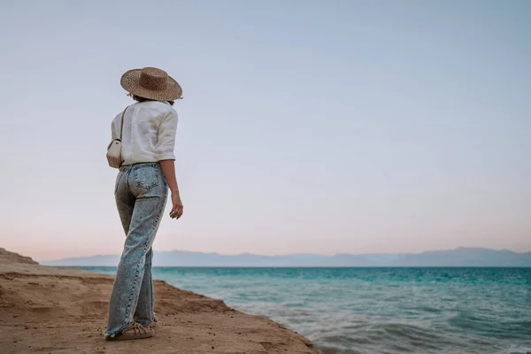 Незнайома жінка стоїть на березі біля Середземного Іонічного моря. Леді турист у солом'яному капелюсі спостерігає за чудовою блакитною поверхнею води на самоті, природним тлом. Вітряна погода, золота година.. — стокове фото