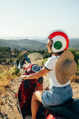Retro scooter 'da oturan ya da kırmızı renkli motosikletli bir kadın, adada dağlarda seyahat ediyor. Miğferli turist harika bir tatil yerinin tadını çıkarıyor. Yaz manzarası.