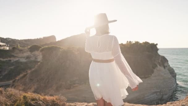Elegante donna in abito bianco arioso a piedi sulla spiaggia scogliera vicino al mare Mediterraneo. Lady godendo esplorare la natura selvaggia, vacanze estive in Grecia. Abito da sposa Boho, sguardo sposa. — Video Stock