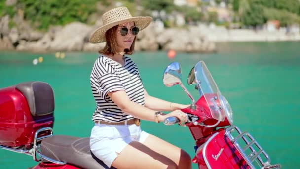 Corfú, Grecia - septiembre de 2021. Mujer sentada en moto Vespa moto en color rojo manzana caramelo, muelle en el pueblo de Paleokastritsa, complejo de lujo. Increíble fondo de agua de mar turquesa. — Vídeo de stock