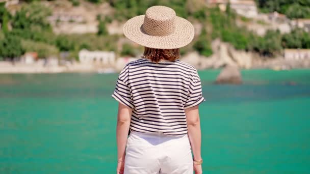 Hermosa escena de verano caliente mujer en camiseta a rayas y sombrero de pie sobre fondo de mar turquesa. Chica en el muelle en Grecia o Italia. Concepto de resort de lujo, viajar — Vídeo de stock