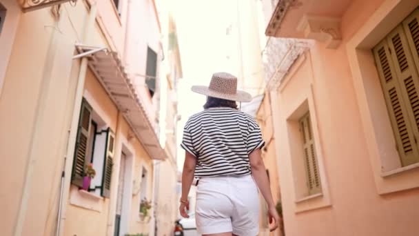Back view of tourist woman in hat exploring old european city on Greece island. Lady sightseeing local Mediterranean architecture, culture. She is having good time wandering around town in summer — Stock Video
