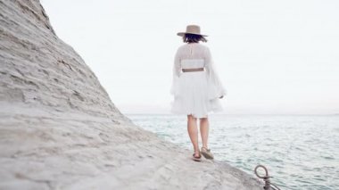 Akdeniz kıyısındaki kil plajda yürüyen beyaz elbiseli yabancı bir kadın. Yunanistan 'da vahşi doğayı keşfeden kadın, yaz tatili.