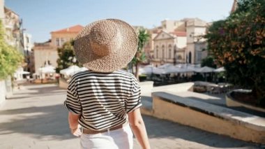 Hasır şapkalı genç bir kadın palmiye ağaçlarıyla sokakta dolaşıyor. Yunan ya da İtalyan tatil köyünde tatil yapan bir kız. Popüler adayı ziyaret eden kadın, seyahat yaşam tarzı konsepti.