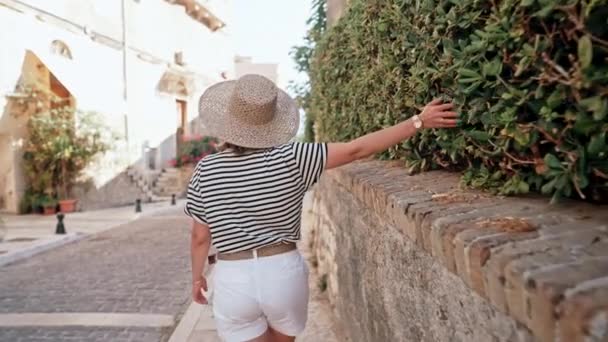 Туристка в соломенной шляпе ходит по узким старинным улицам. Девушка проводит отпуск в Греции. Осмотр достопримечательностей местной архитектуры. Она хорошо проводит время в городе летом. — стоковое видео