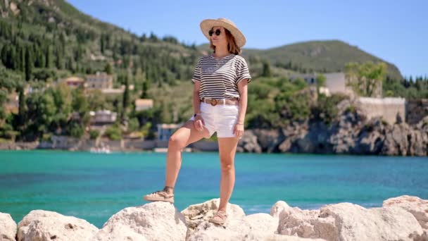 Bella scena estiva calda donna in t-shirt a righe e cappello in piedi su sfondo turchese mare e montagne. Ragazza sulla spiaggia di pietra in Grecia. Concetto resort di lusso, viaggiare — Video Stock