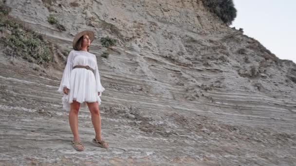 Beyaz elbiseli genç bir kadın portresi, ipe bağlı sandaletler ve çamur plajında poz veren hasır şapka. Yunanistan 'da yaz tatilinin tadını çıkaran şık model bayan. Boho düğün kıyafeti, modern gelin görünüşü. — Stok video