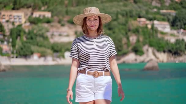 Sommarscen - leende kvinna i halmhatt som går mot kameran. Hav, motorbåt på bakgrunden. Flicka på piren i Grekland eller Italien. Lyx resort koncept, resa — Stockvideo