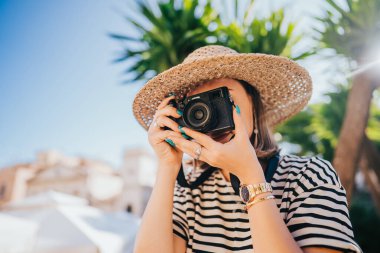 Eski Yunan şehrinde palmiye ağacının altında duran turist kadın fotoğrafçı. Yaz tatili kavramı. Kadın adayı keşfediyor. Küçük dijital kamerayla fotoğraf çeken amatör..