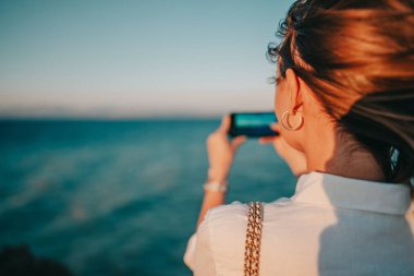 Mavi deniz kıyısındaki sahilde cep telefonuyla video çeken genç bir kadın. Akıllı telefon kullanan, sosyal uygulama için fotoğraf çeken, tatilden zevk alan bir kız..