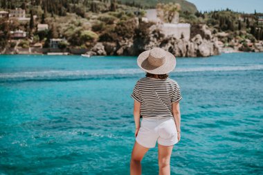 Çizgili gezi kıyafeti giymiş, hasır şapkalı bir kadın. Taş plajda duran bir kadın. Teal denizi arka planı. Güney Avrupa Riviera, seyahat yeri, lüks tatil konsepti.