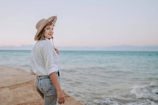Красуня стоїть на березі моря. Леді турист у солом'яному капелюсі спостерігає прекрасну блакитну поверхню води, природне тло. Вітряна погода, золота година.. — стокове фото