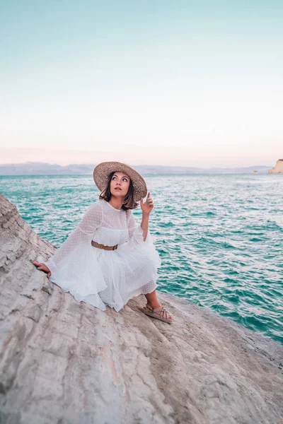 Portret młodej kobiety w białej sukience, modne sandały i słomkowy kapelusz na glinianej plaży. Stylowa modelka ciesząca się wakacjami w Grecji. Boho strój ślubny, nowoczesny wygląd panny młodej. — Zdjęcie stockowe
