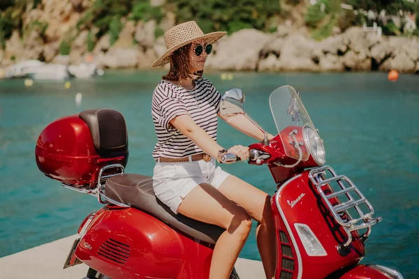Korfu, Grecja - wrzesień 2021. Kobieta siedząca na skuterze motocyklowym Vespa w kolorze jabłkowego czerwonego cukierka, molo w wiosce Paleokastritsa, luksusowy ośrodek. Niesamowite turkusowe tło wody morskiej. — Zdjęcie stockowe