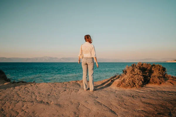 Невпізнавана жінка стоїть біля скелі над середземноморським морем. Леді дивиться на прекрасну блакитну поверхню води сама, природне тло. Вітряна погода, золота година.. — стокове фото