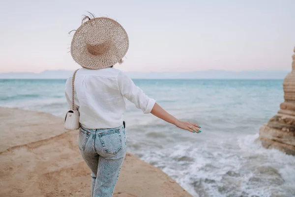 Csinos nő áll a tengerparton közel a Földközi-tengerhez. Lady turista szalmakalapban nézi gyönyörű kék víz felszínét, a természet hátterét. Szeles idő, aranyóra. — Stock Fotó