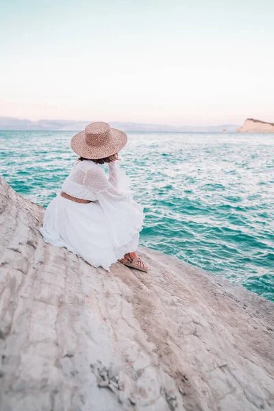 Portré felismerhetetlen nő fehér boho ruha, kötél divatos szandál és szalma kalap ül egyedül agyag strandon. Stílusos modell hölgy élvezi nyári vakáció Görögországban. — Stock Fotó