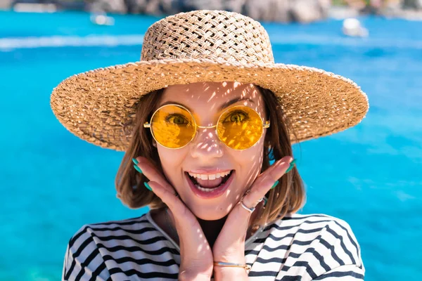 Surprise jolie femme en lunettes de soleil jaunes et chapeau de paille dit WOW avec bouche ouverte étonné de la caméra sur fond bleu sarcelle mer. Dame sur la destination de voyage de luxe, concept de style de vie vacances. — Photo