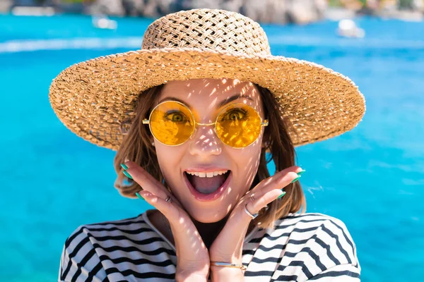 Sorpresa bella donna in occhiali da sole gialli e cappello di paglia dice WOW con la bocca aperta in stupore per la fotocamera su sfondo blu acqua. Lady sulla destinazione di viaggio di lusso, concetto di stile di vita di vacanza. — Foto Stock