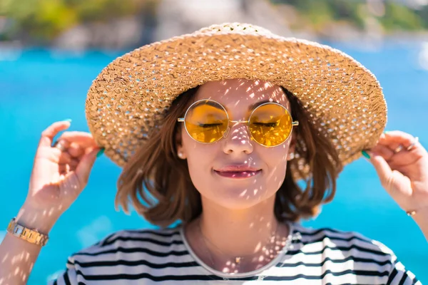 Jolie femme aux lunettes de soleil jaunes et chapeau de paille souriant sincèrement à la caméra sur fond de mer bleu sarcelle. Dame sur la destination de voyage de luxe, concept de style de vie vacances. — Photo