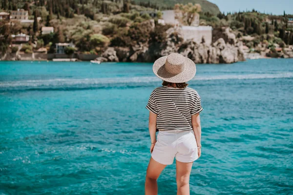 Visszapillantás a csíkos tengerjáró ruhás nőre, szalmakalapban. Egy hölgy áll a Stone Beach-en. Teal tengeri háttér. Dél-Európa Riviéra, utazási cél, luxus nyaralás koncepció. — Stock Fotó