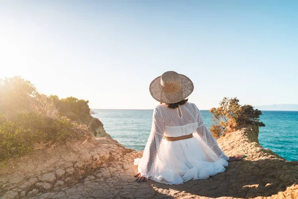 Młoda stylowa kobieta siedząca na klifie nad Morzem Śródziemnym, chłodząca się. Pani w sukience i słomkowym kapeluszu ciesząca się wakacjami na tropikalnej wyspie. Tło dzikiej przyrody. — Zdjęcie stockowe