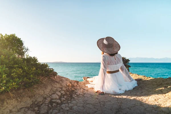Boho turystka siedzi na klifie nad Morzem Śródziemnym Canal DAmour, chłodzenie. Pani w sukience i słomkowym kapeluszu ciesząca się wakacjami na Korfu, Grecja. Tło dzikiej przyrody. — Zdjęcie stockowe