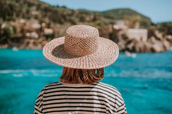 Widok z tyłu kobiety w stroju rejsowym w paski, słomiany kapelusz. Kobieta stojąca na kamiennej plaży. Teal tło morza. Riwiera Europa Południowa, cel podróży, koncepcja luksusowych wakacji. — Zdjęcie stockowe