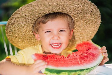 Küçük tatlı bir çocuk, sulu karpuz yerken doğal yeşil arka planda oturuyor. Hasır şapkalı sevimli çocuk gülümsemeli taze kavun meyvesinin tadını çıkarıyor..