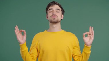 Calm man relaxing, meditating, refuses stress. Brown haired guy breathes deeply, calms down green studio background. Yoga, moral balance, zen concept. High quality 4k