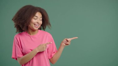 Gülümseyen öğrenci reklam için boş bir yer arıyor. Önerilen kadın fotokopi alanını işaret ediyor. Kadın, yeşil arka planda izole edilmiş bir şey sunuyor. Yüksek kalite 4k