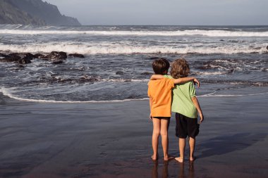 İki genç çocuk, kardeşler ya da arkadaşlar, plajda siyah volkanik kumla, Atlantik Okyanusu 'nda birbirlerine sarılıyorlar. Tanınmayan çocuklar, arkadaşlık, aile bağı, çocukluk, İspanya 'da seyahat, Kanarya Adası