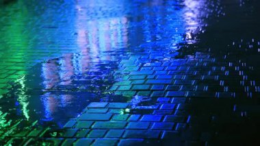 Gece kaldırım taşı sokağındaki su birikintisine düşen yağmur damlaları mavi ve yeşil şehir ışıklarıyla aydınlanıyor. Yansımalar ve dalgalanmalar karamsar, fütüristik titreşimlerle sinematik bir şehir atmosferi yaratır..