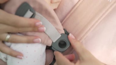 Anne çocuğunu tutarken bebek taşıyıcısının tokasını bağlıyor. Ebeveyn bakımı, güvenlik, bağlanma ve ebeveynlik rutinine odaklan. Koruma, çocuk bakımı ve güvenli bebek giyimi kavramı.