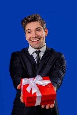 Olgun Takım Elbiseli İş Adamı, Kameraya Hediye Kutusu Veriyor. Adam mutlu, mavi duvarda gülümsüyor. Pozitif ruh hali, tatil, ikramiye, doğum günü konsepti. Yüksek kalite
