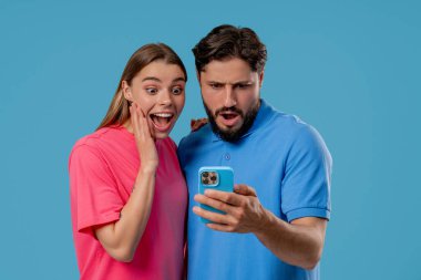 Cep telefonu kullanan genç bir çift. Kadın ve erkek izliyor, konuşuyor, gülümsüyor, akıllı telefonla gülüyor. Sosyal uygulamalar, ağlar, teknoloji konsepti. Mavi stüdyo arka planı.