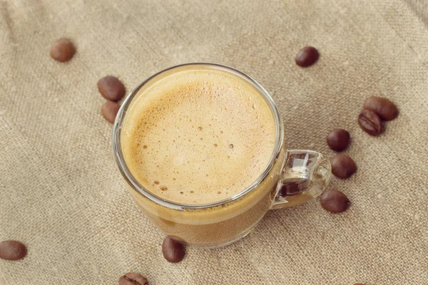Cappuccino com crema em uma xícara transparente em saquear. grãos de café fechar — Fotografia de Stock