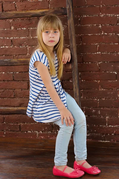 Niña de 6 años en jeans y chaleco se sienta en una escalera cerca de la pared — Foto de Stock