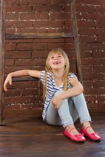 Menina 6 anos de idade em jeans e um colete senta-se no chão ao lado da parede de tijolo. cheio de risos — Fotografia de Stock