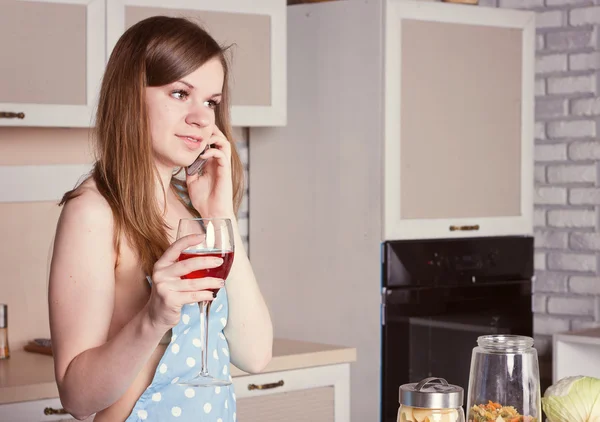 Menina na cozinha vestindo um avental com um copo de vinho no telefone — Fotografia de Stock