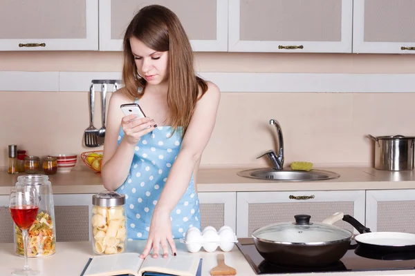 Meisje in de keuken het dragen van een schort over zijn naakt lichaam bereidt en kijkt naar telefoon — Stockfoto