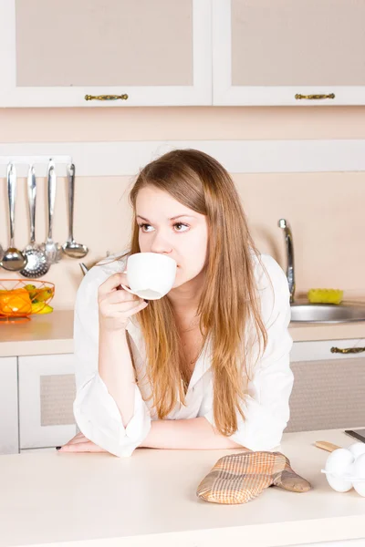 Ragazza una camicia bianca da uomo con lunghi capelli fluenti sta bevendo tè gomiti della cucina sul tavolo. mattina — Foto Stock
