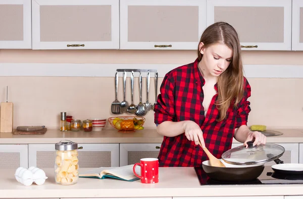 Se prepara la muchacha con los cabellos largos que fluyen en la camisa roja masculina. la cocina —  Fotos de Stock