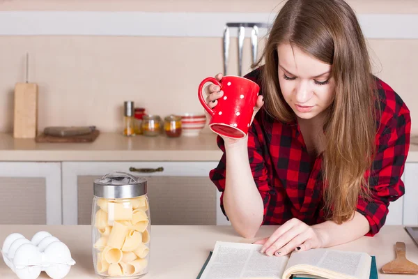 Ragazza in camicia rossa che beve tè e legge. in cucina. Orizzonte — Foto Stock