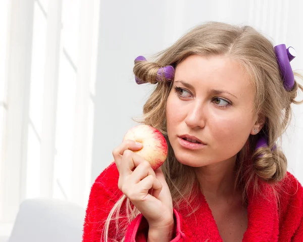 Girl in red robe and curlers eating an apple — Stock Photo, Image